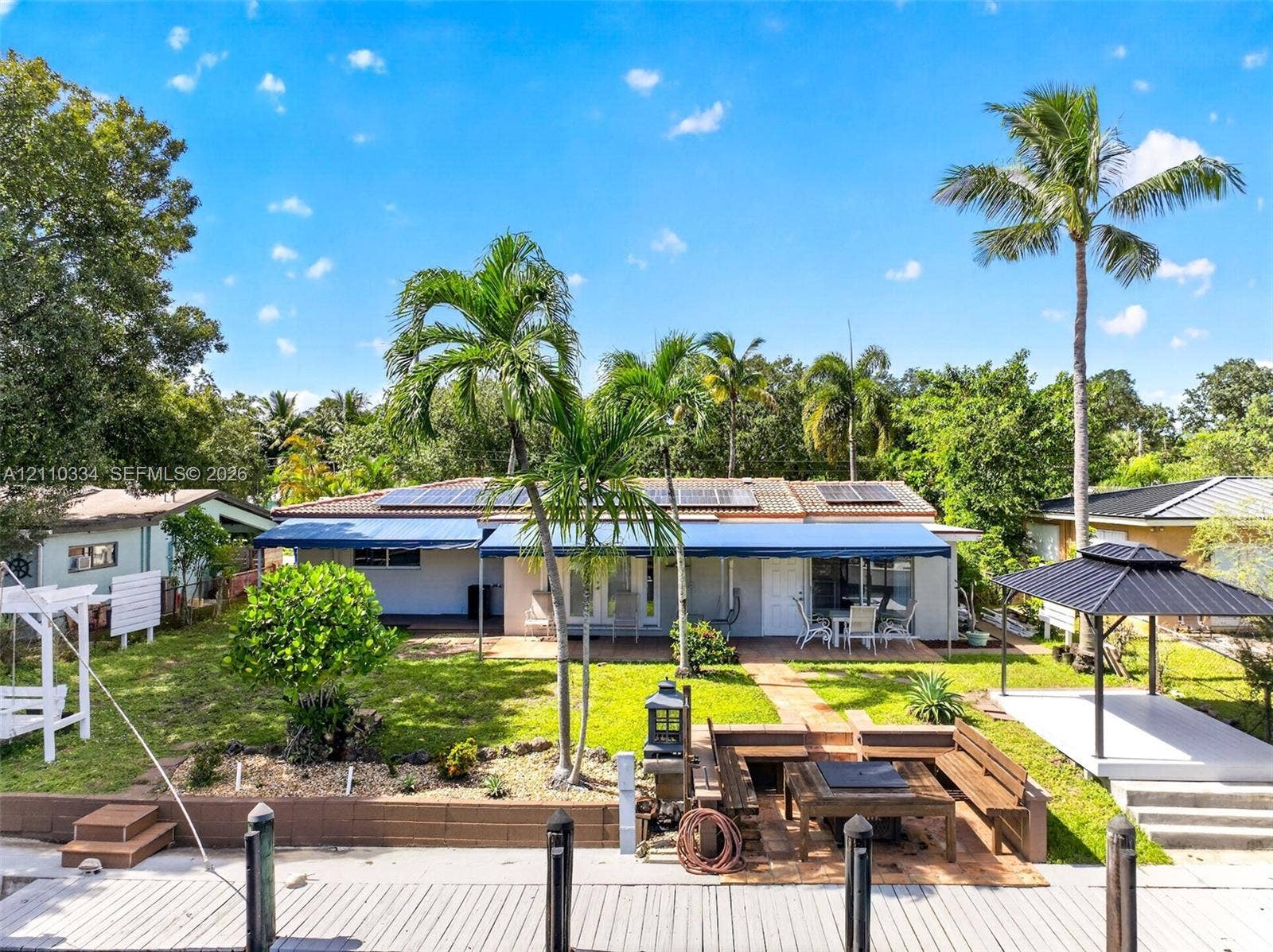 2100 SW 23rd Terrace, Fort Lauderdale, FL 33312 Waterfront backyard featuring a tiered wooden bulkhead, integrated fire pit seating area, covered patio with retractable awnings, detached gazebo, and roof-mounted solar panels