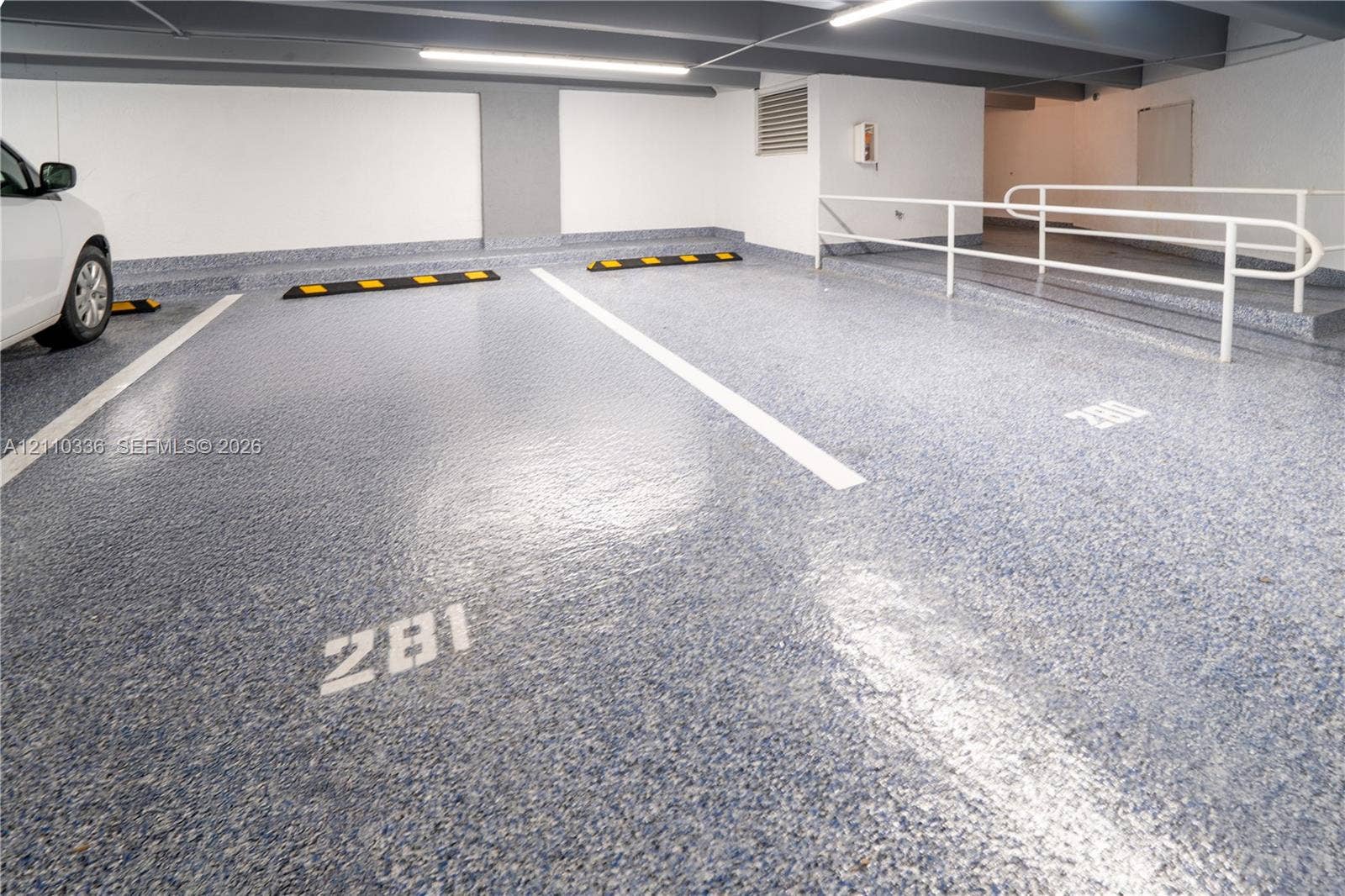 16445 Collins Avenue, #WS-7B, Sunny Isles Beach, Sunny Isles Beach, FL 33160 Covered parking garage featuring high-gloss speckled epoxy flooring, painted concrete walls, overhead strip lighting, and a white metal safety railing