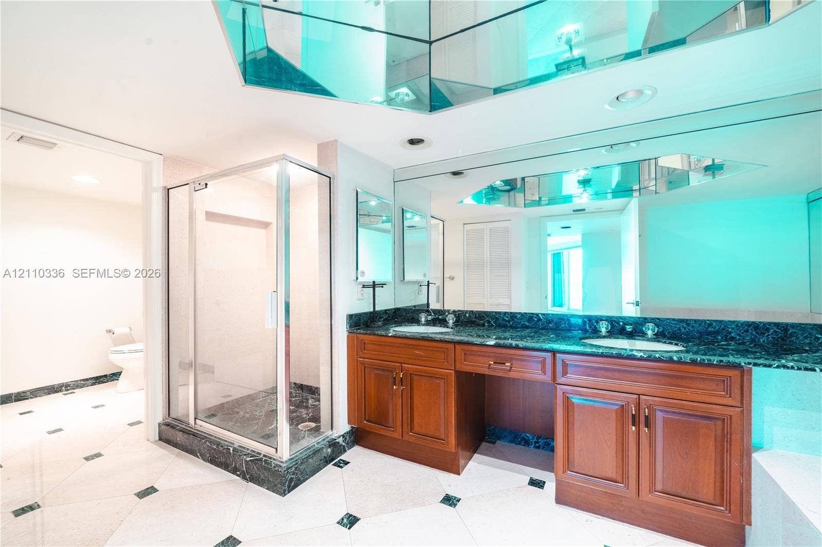 16445 Collins Avenue, #WS-7B, Sunny Isles Beach, Sunny Isles Beach, FL 33160 Double vanity with stained wood cabinetry and stone countertops, glass-enclosed shower, expansive wall-to-wall mirror with faceted mirrored accents, and tiled flooring with decorative stone inlays