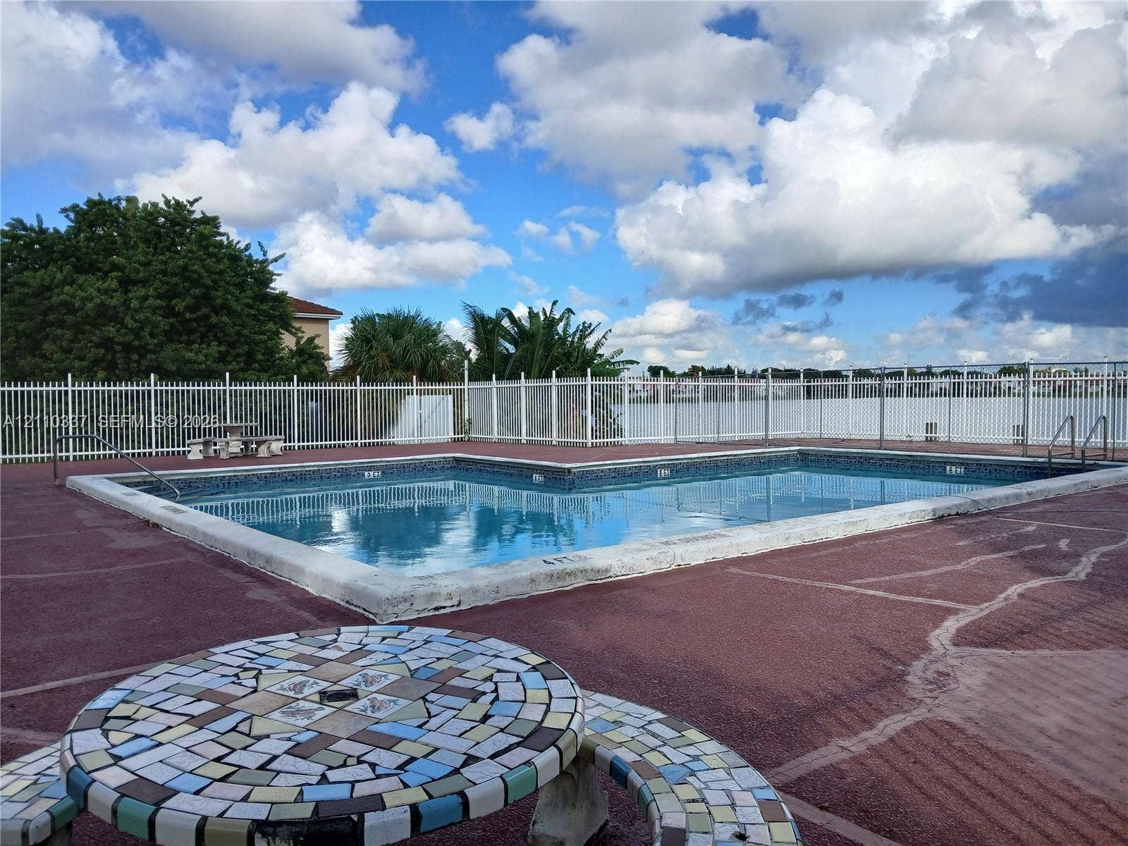 In-ground swimming pool with tile border, surrounded by a paved deck and enclosed by a white metal perimeter fence
