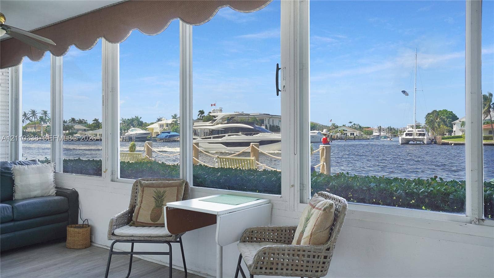 Enclosed sunroom featuring expansive floor-to-ceiling windows, wood-finish flooring, and a retractable fabric awning, overlooking a private canal with bulkhead and nautical access