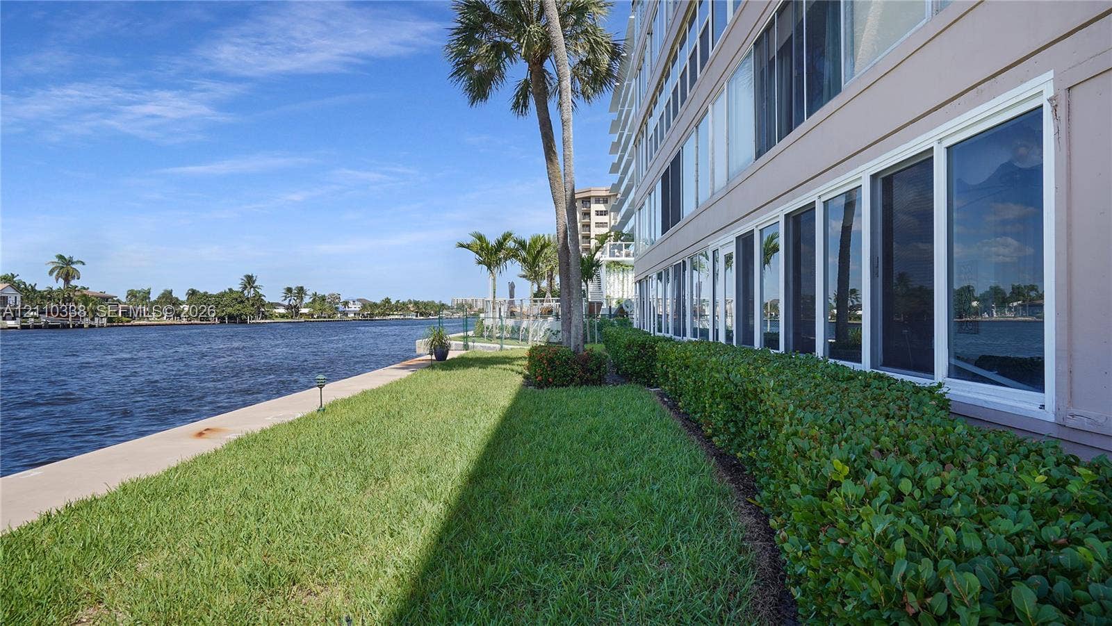 Waterfront grounds featuring a concrete seawall, manicured lawn, perimeter hedge landscaping, and palm trees