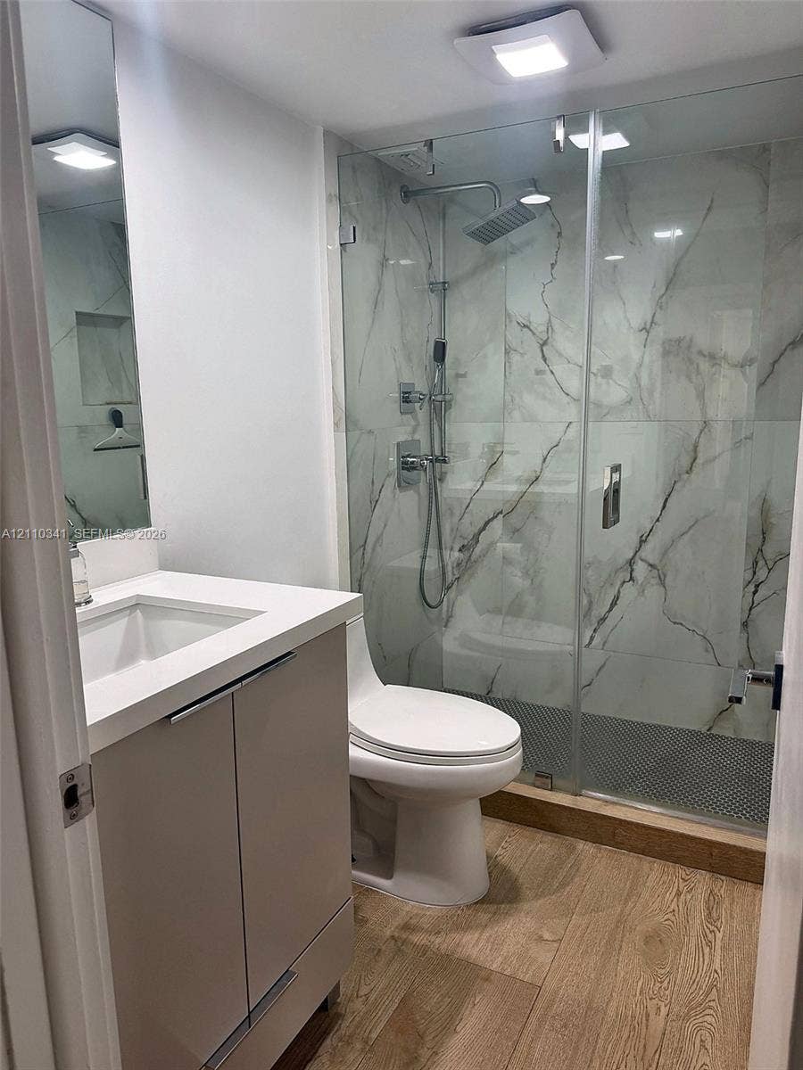 Bathroom featuring a vanity with a white countertop and rectangular sink, frameless glass-enclosed shower with large-format stone-patterned wall tile, rain-style shower head, and wood-finish flooring