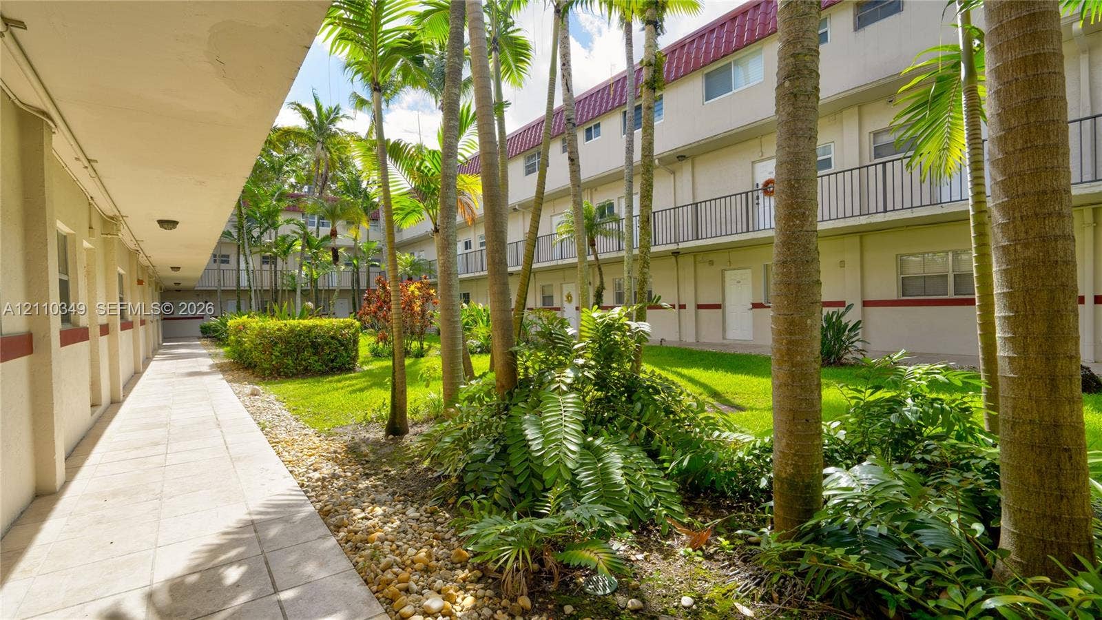 8600 SW 67th Avenue, #905, Pinecrest, Pinecrest, FL 33156 Covered exterior walkway featuring a tiled floor, manicured lawn, stone ground cover, mature palm trees, and multi-level railings