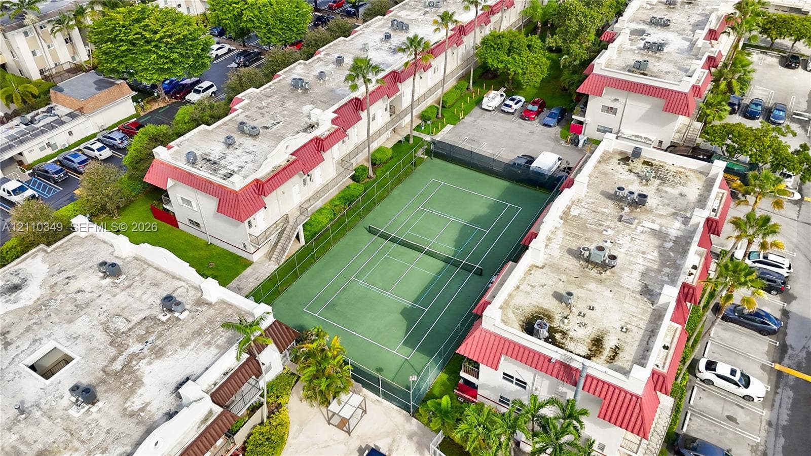 8600 SW 67th Avenue, #905, Pinecrest, Pinecrest, FL 33156 Aerial perspective of a multi-unit property featuring a fenced tennis court, red metal roofing, and surrounding palm trees