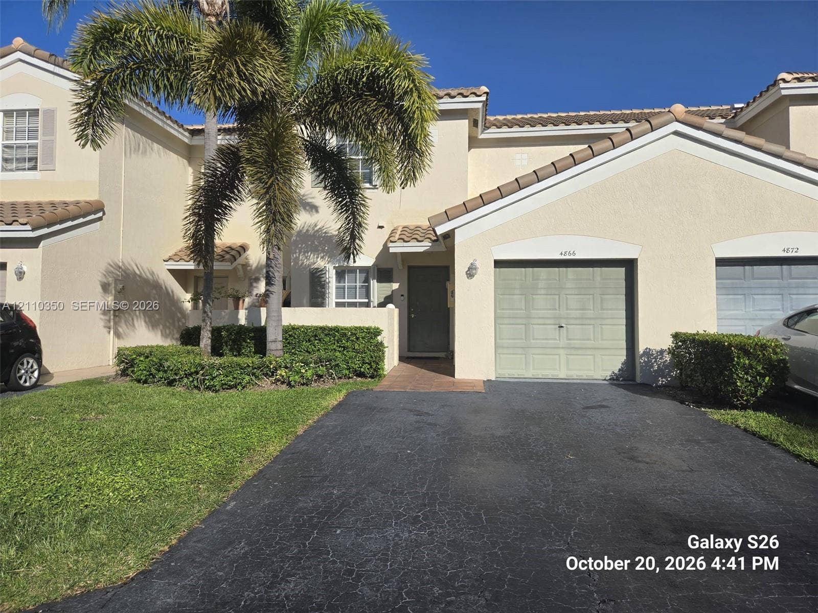 Stucco exterior featuring a tile roof, single-car garage with a paneled door, private entryway, and mature palm tree landscaping
