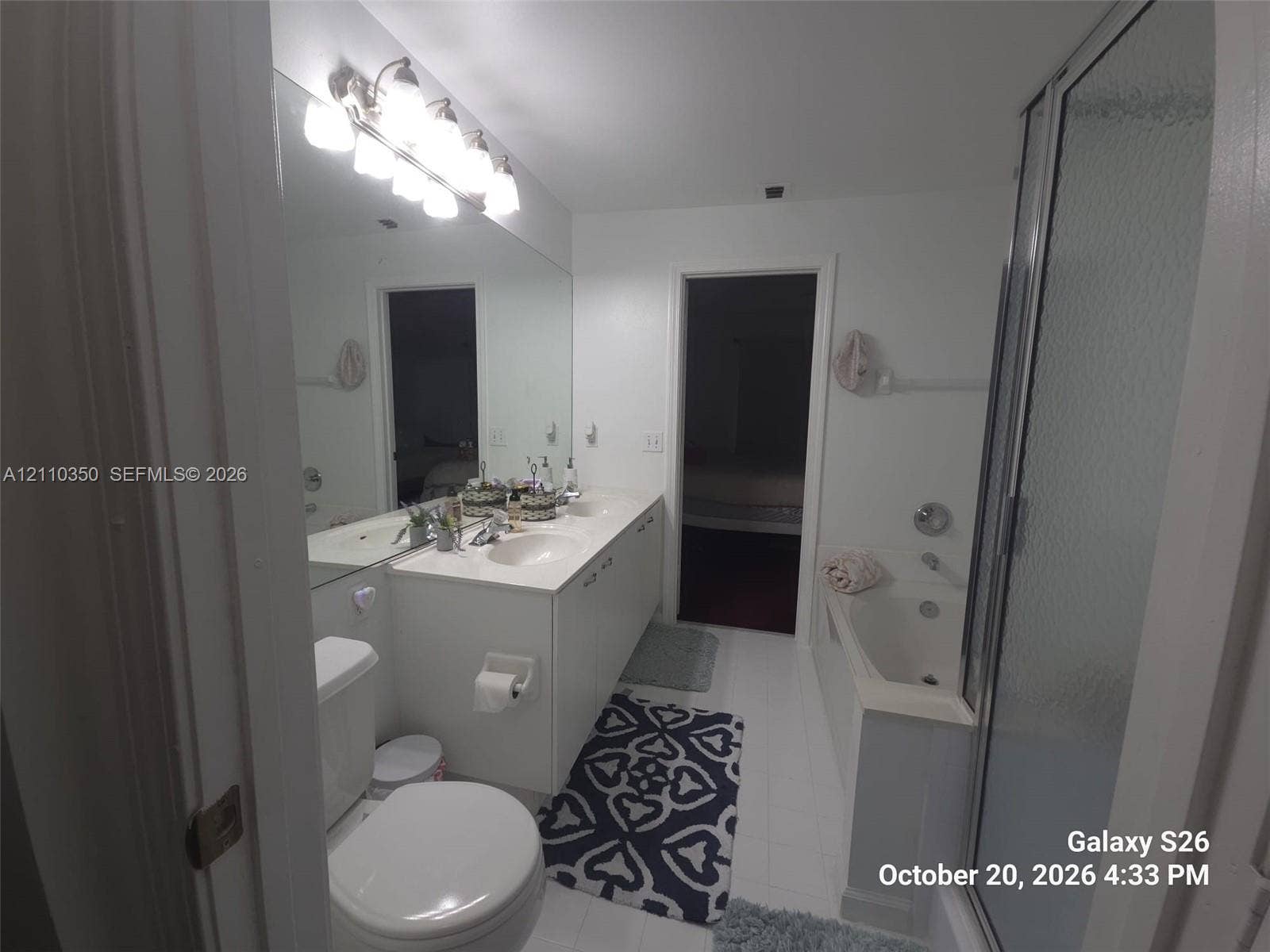 Bathroom featuring a dual-sink vanity with solid-surface countertop, full-width vanity mirror, multi-bulb wall-mounted lighting fixture, combination soaking tub and shower enclosure, and tiled flooring