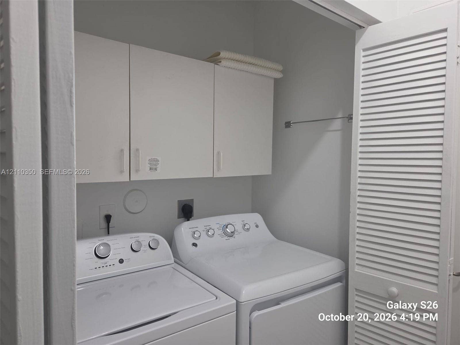 Laundry alcove featuring white louvered doors, upper wall-mounted cabinetry, and a horizontal metal hanging rod