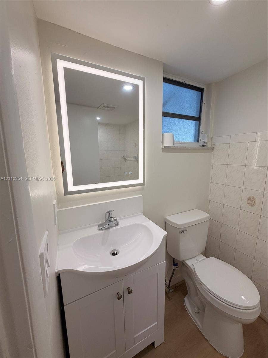 8801 W Flagler Street, #309, Miami, Miami, FL 33174 White vanity cabinet with an integrated basin, wall-mounted rectangular LED mirror, two-piece toilet, and tile wall detailing