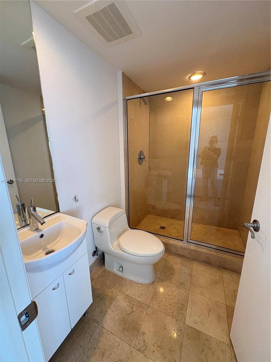 Bathroom featuring stone-tile flooring, a glass-enclosed shower with tiled surround, a white vanity cabinet, a wall-mounted mirror, and a one-piece toilet