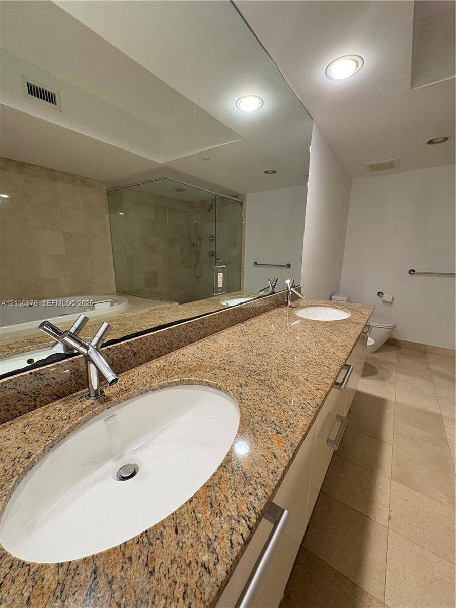 Dual-basin vanity with stone countertops, integrated oval sinks, polished metal fixtures, full-width mirror, and a glass-enclosed shower