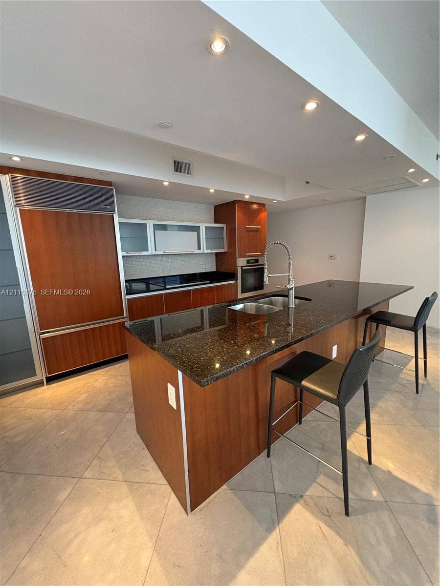 Kitchen featuring wood cabinetry, integrated refrigerator, dark stone countertops, central island with double-basin sink, and tile flooring