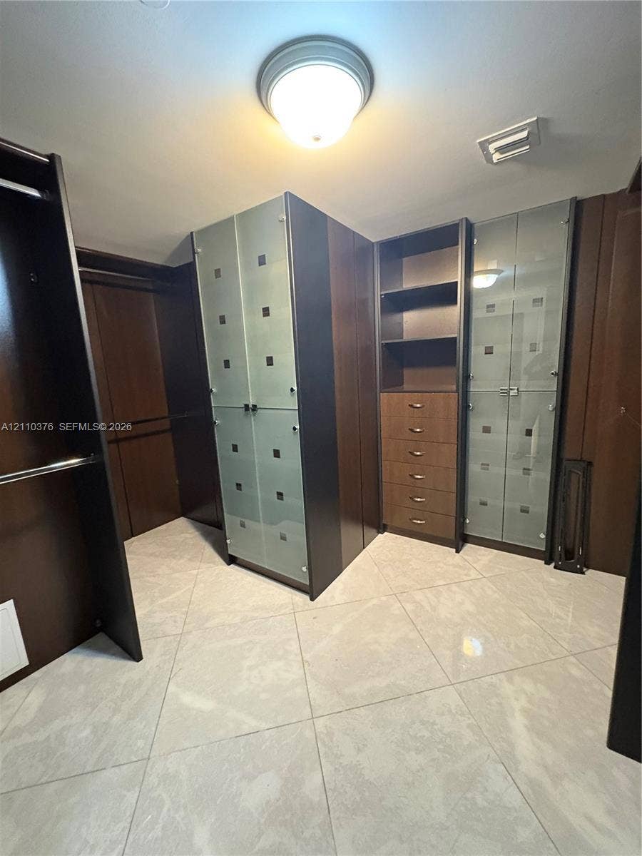 Custom storage area featuring floor-to-ceiling cabinetry with dark wood-finish, integrated glass-front doors, open shelving, and six pull-out drawers, finished with large-format light-toned tile flooring