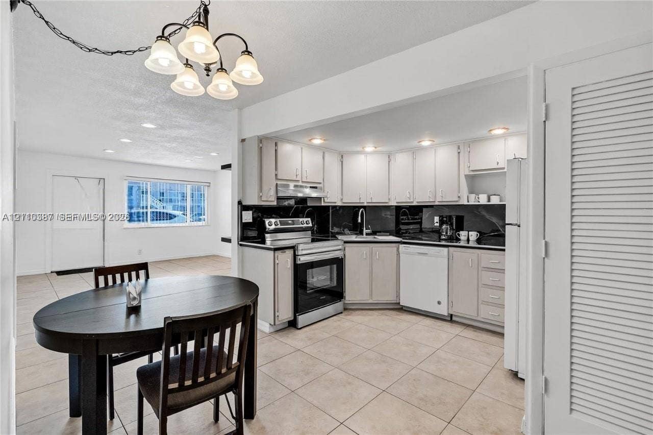 Kitchen featuring white shaker-style cabinetry, black stone countertops, stainless steel appliances, and ceramic tile flooring, complemented by a multi-light chandelier