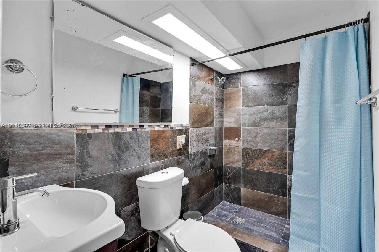 Bathroom featuring large-format multicolored stone wall tiling, a pedestal sink with a polished chrome faucet, a wall-mounted mirror, and a walk-in shower area with recessed lighting