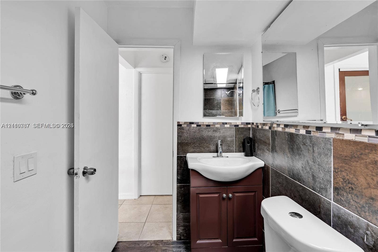 Bathroom featuring a dark wood-finish vanity with a ceramic basin, chrome faucet, expansive wall-to-wall mirror, and floor-to-ceiling tilework with mosaic decorative accents
