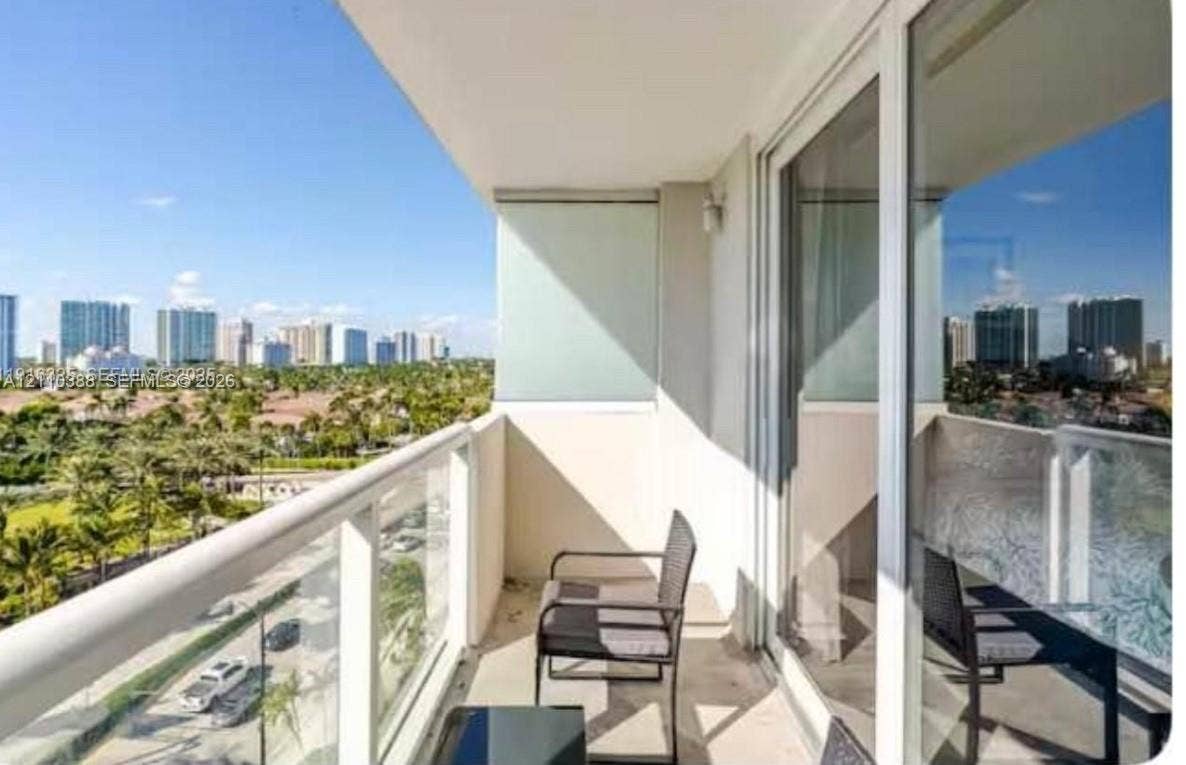 Covered balcony featuring a glass-panel railing, expansive sliding glass doors, and a panoramic perspective of the cityscape and surrounding greenery