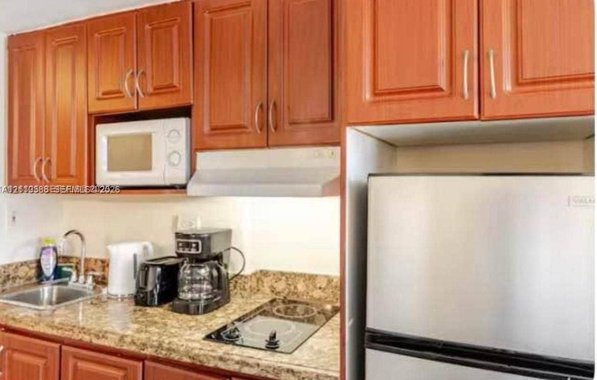 Kitchen featuring wood-toned cabinetry, stone-patterned countertops, stainless steel sink with a high-arch faucet, integrated stovetop, and built-in microwave