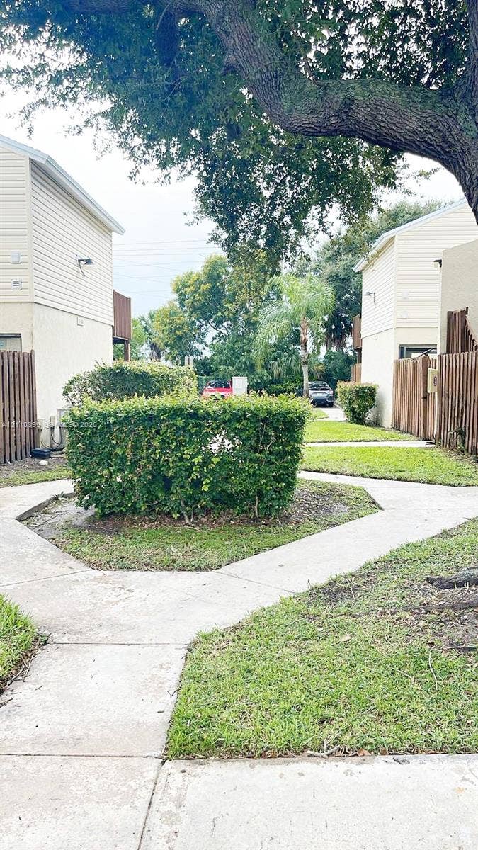 Exterior grounds featuring concrete walkways, manicured landscaping, mature trees, and wooden fencing