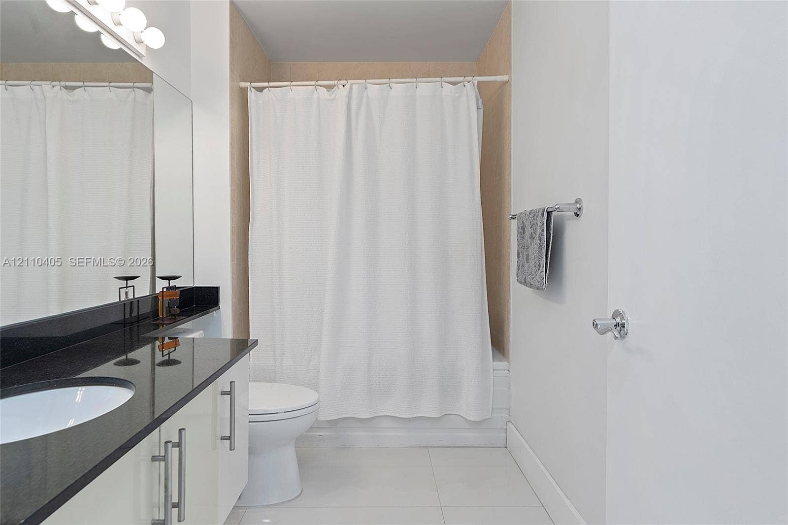 Bathroom featuring a vanity with dark stone countertop, integrated sink, polished metal hardware, full-width mirror with integrated lighting, and a bathtub with tile surround