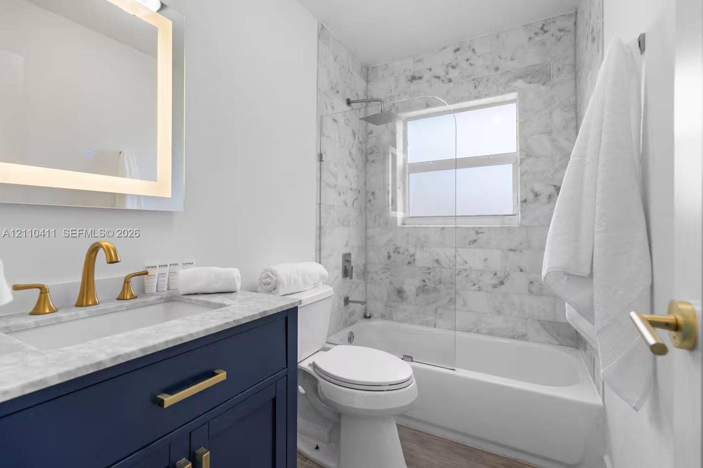 Navy blue vanity with stone-top surface, brushed gold-tone faucet, integrated LED-backlit mirror, stone-tiled shower surround, and glass shower panel