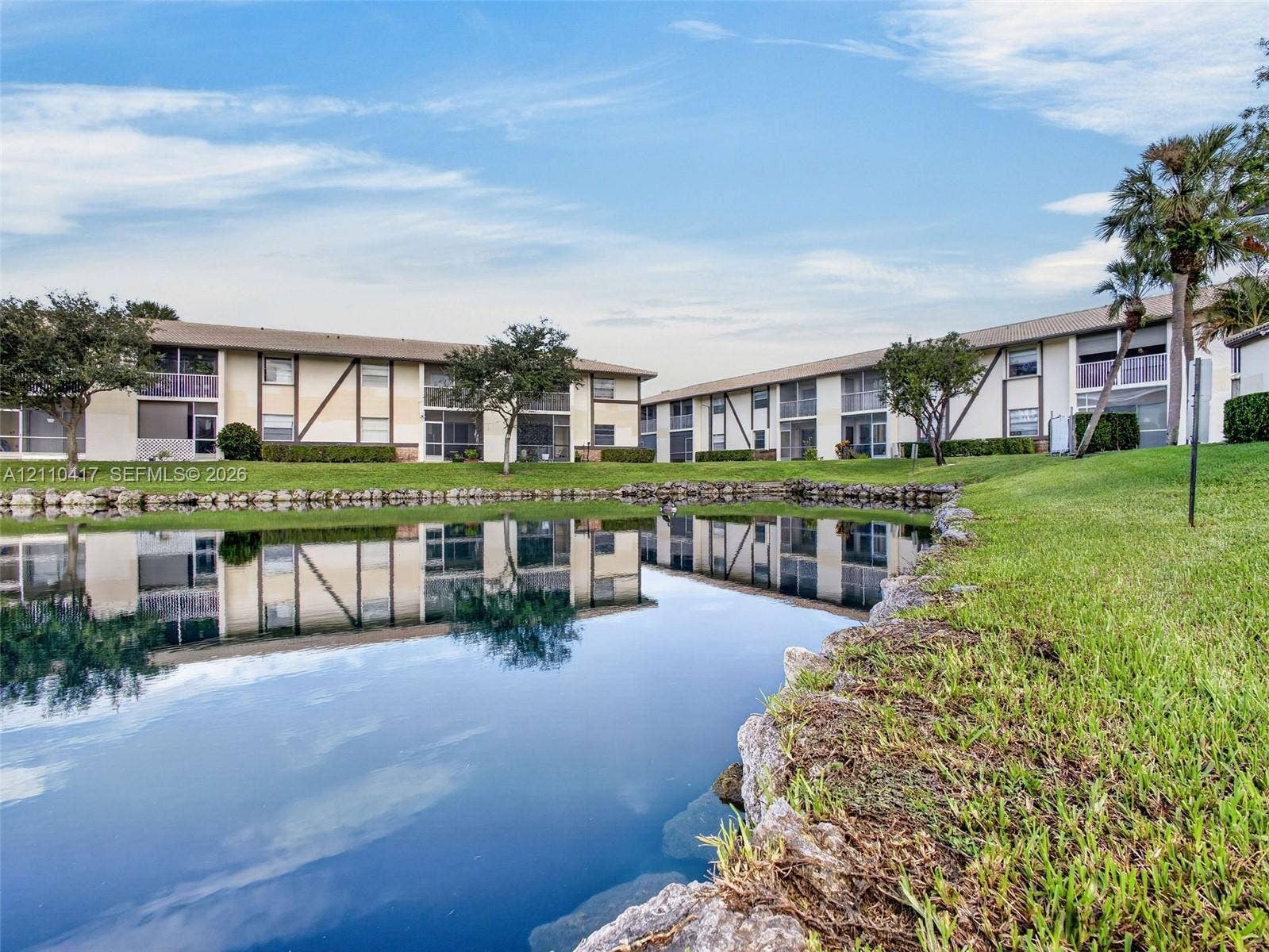 2369 NW 89th Drive, #514, Coral Springs, FL 33065 Waterfront multi-story residential building with stone-lined shore, manicured lawn, and mature palm trees