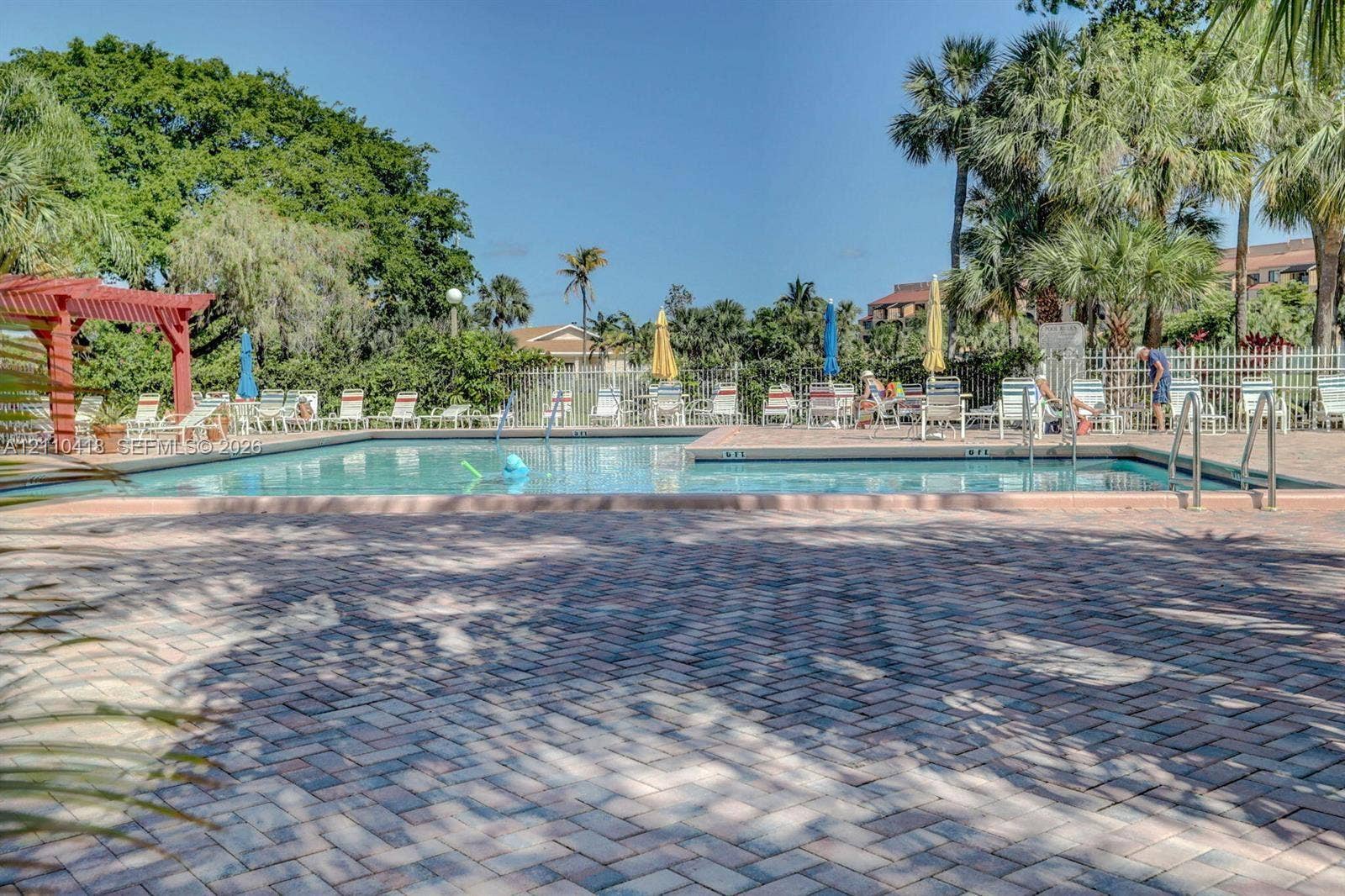 950 Egret Circle, #5409, Delray Beach, Delray Beach, FL 33444 Outdoor swimming pool featuring a brick paver deck, perimeter metal fencing, and a red wood-finish pergola