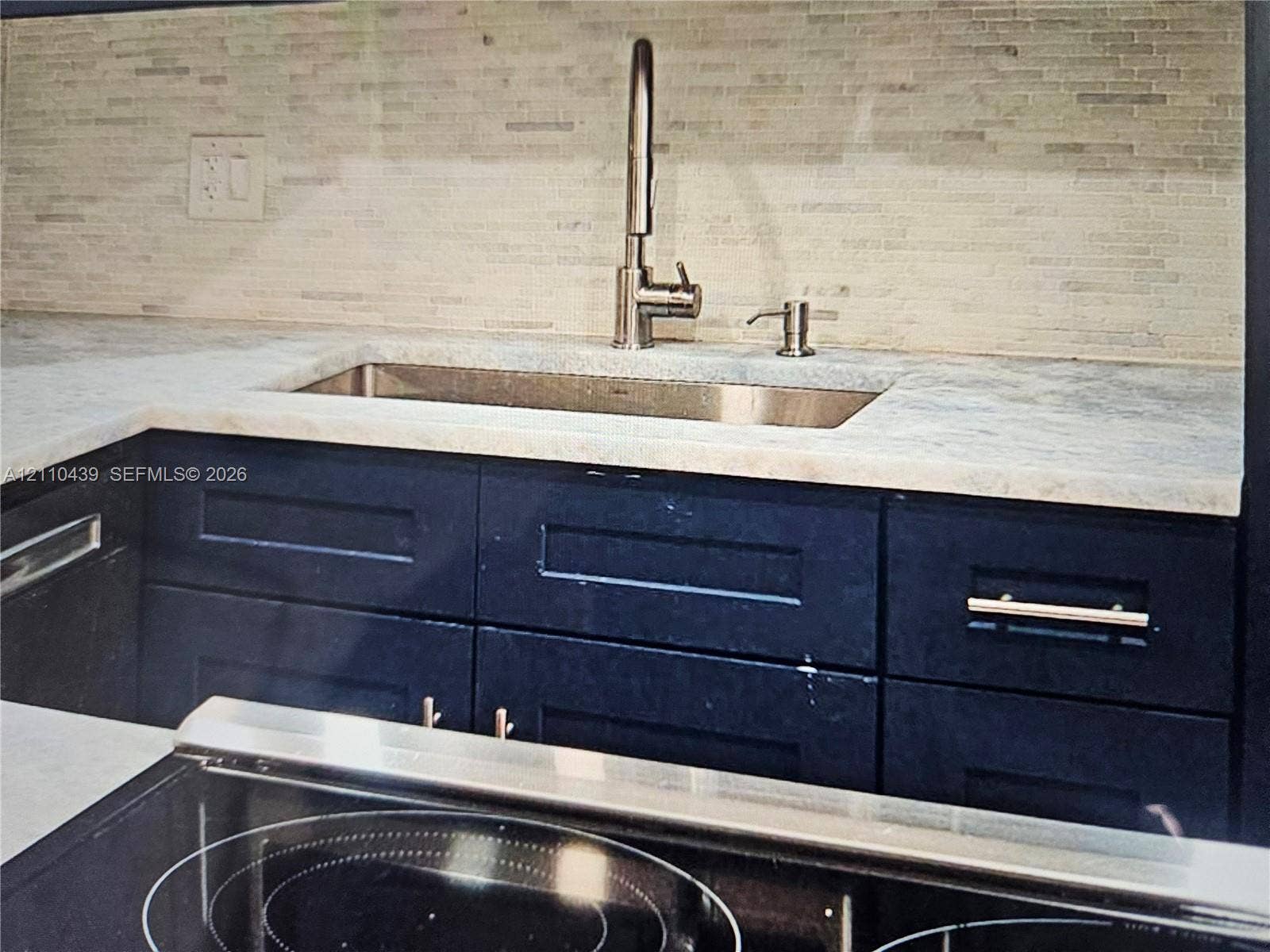 Undermount stainless steel sink with a high-arch pull-down faucet, stone-finish countertops, and linear glass tile backsplash, accented by deep-toned shaker cabinetry and a smooth-surface cooktop
