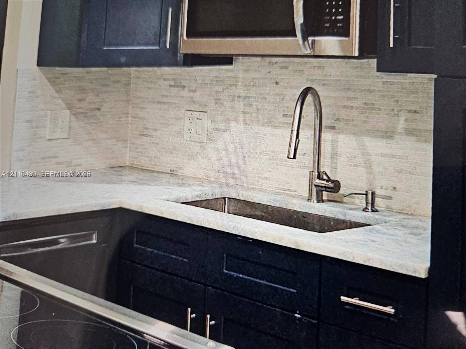 Kitchen featuring dark-toned cabinetry, light-toned stone-finish countertops, linear tile backsplash, stainless steel under-mount sink, and gooseneck pull-down faucet