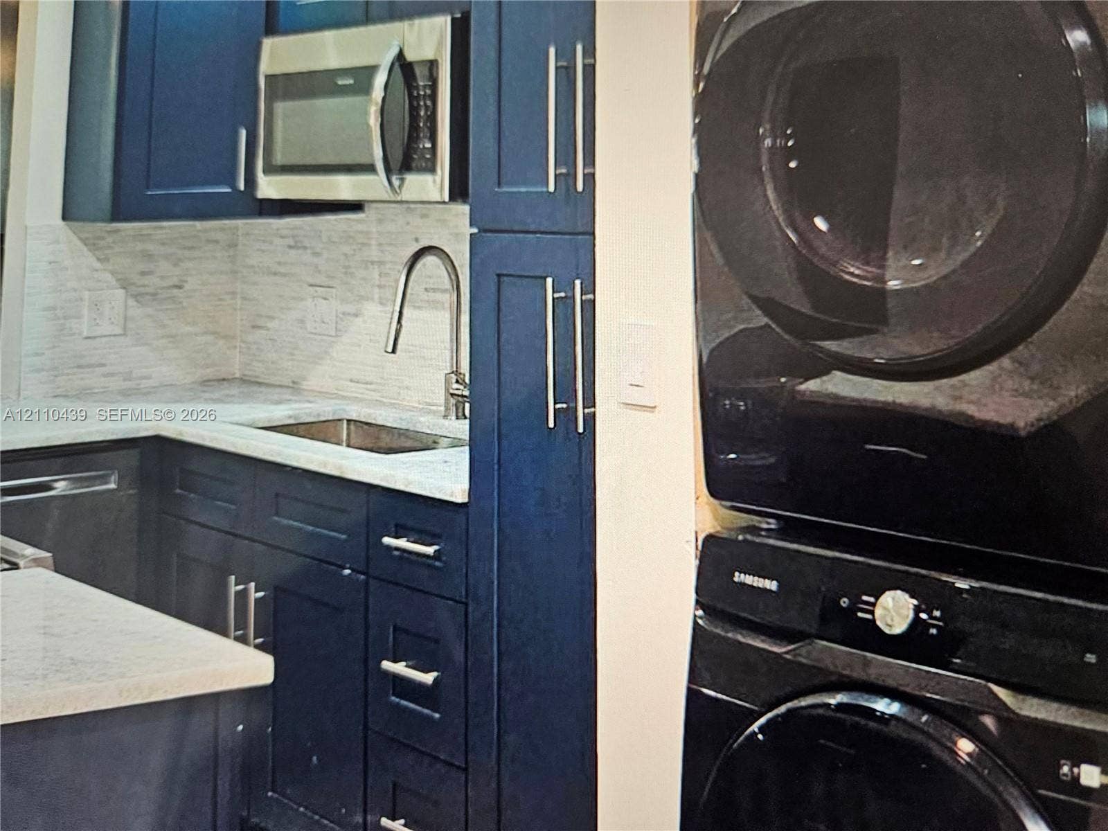 Navy blue shaker-style cabinetry with satin nickel hardware, stone-finish countertops, tiled backsplash, stainless steel under-mount sink with high-arc faucet, and stacked laundry unit