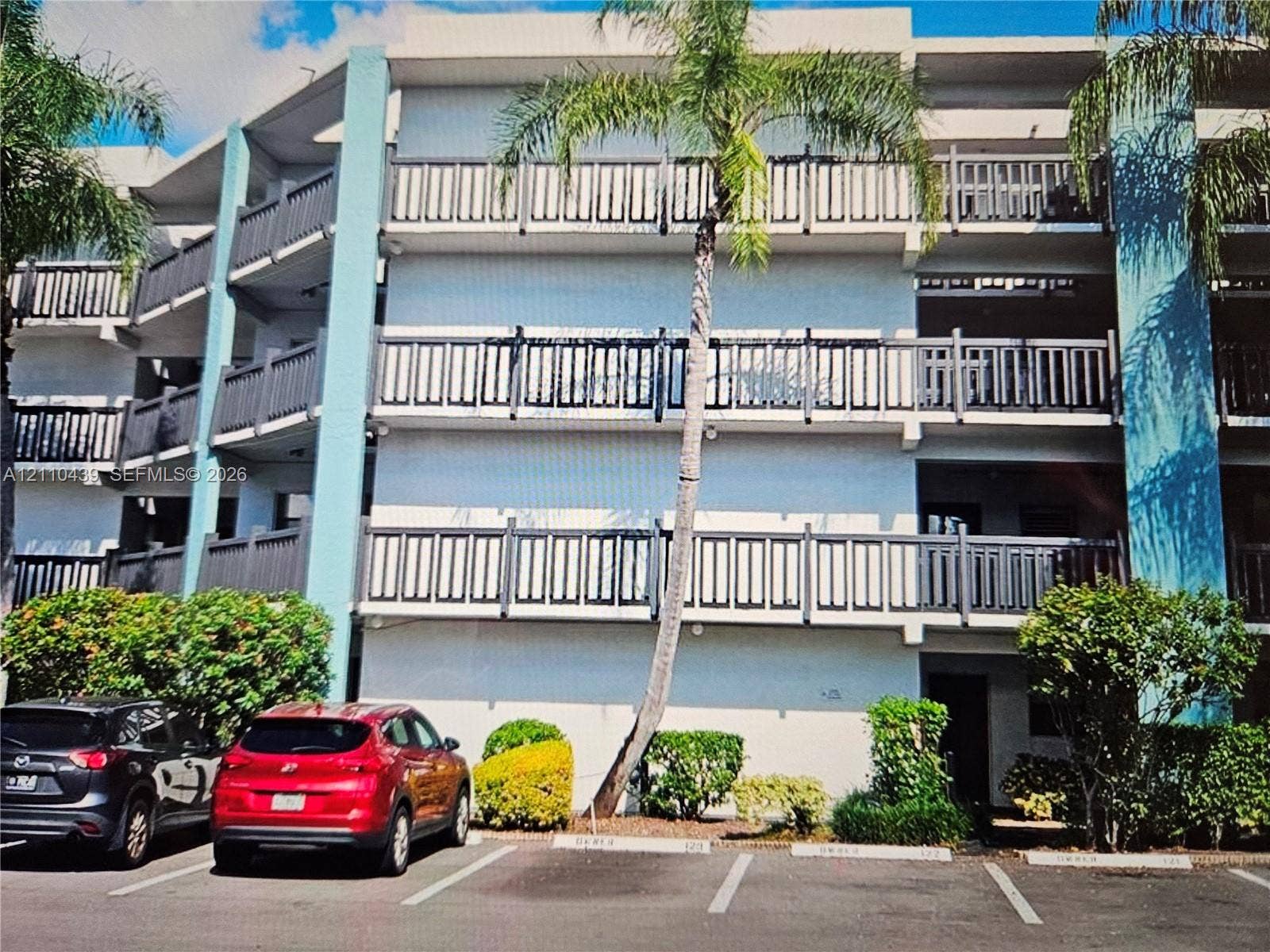 Four-story building exterior featuring vertical turquoise architectural accents, multi-level balconies with dark railings, and paved surface parking