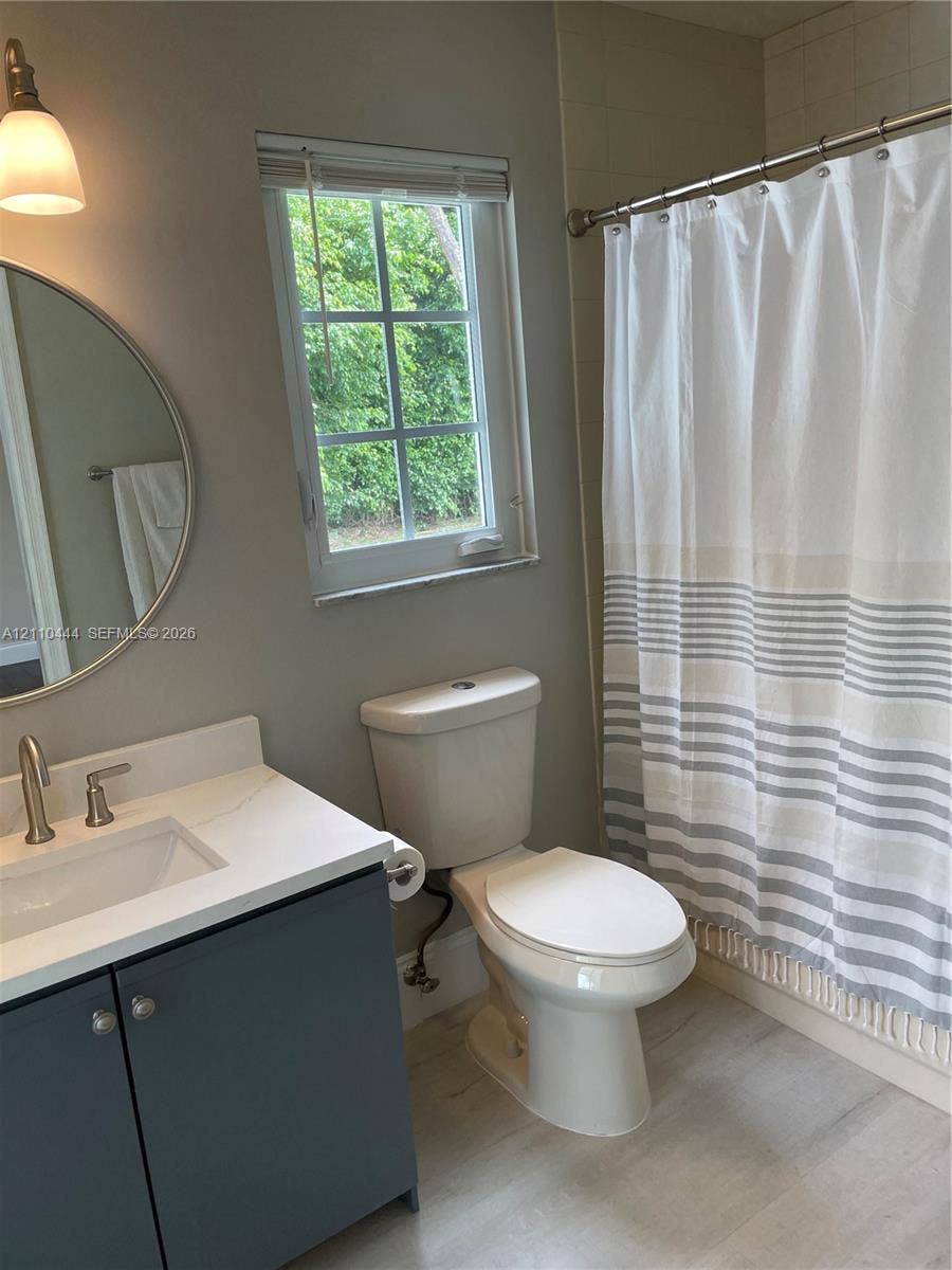 Bathroom featuring a charcoal-toned vanity with a white countertop, circular wall-mounted mirror, single-handle brushed nickel faucet, and wood-finish flooring