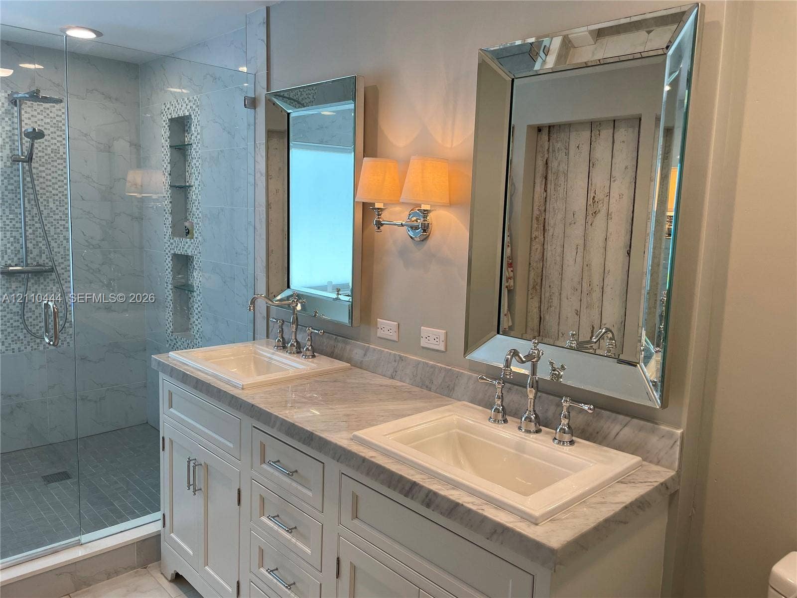 Double vanity featuring stone countertops, integrated rectangular sinks, polished metal widespread faucets, framed mirrors, and a walk-in shower with stone tile surround and built-in recessed shelving