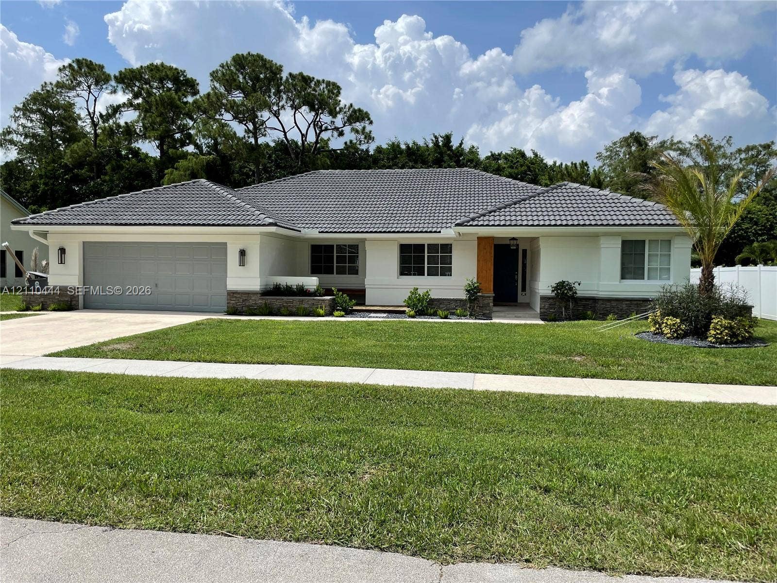 Single-story residence featuring a gray tiled roof, light-colored stucco exterior, stone masonry accents, a double-car garage, and a manicured lawn