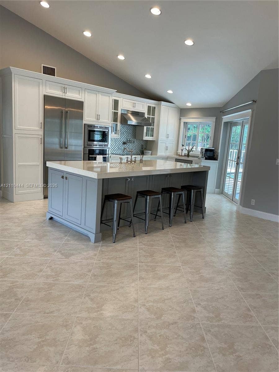 Kitchen featuring an oversized island with quartz countertops, custom cabinetry, stainless steel appliances, tile flooring, and vaulted ceilings with recessed lighting
