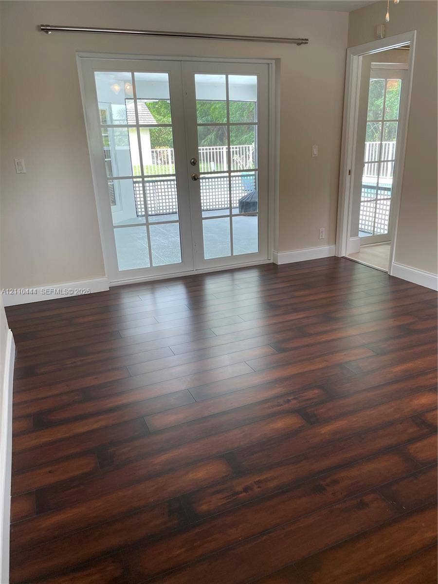 Room with dark wood-finish flooring, white-trimmed French doors with glass panes, and a separate doorway featuring a matching glass-paned door