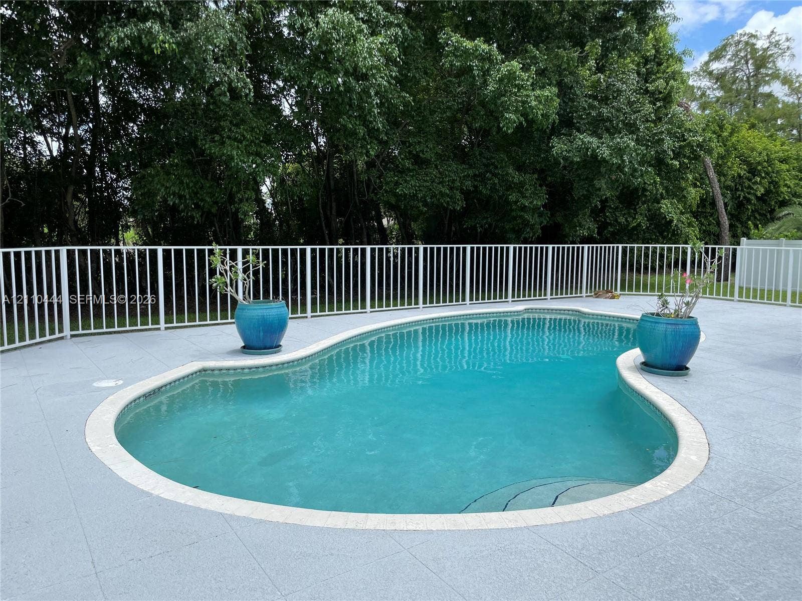 In-ground pool with stone coping, surrounded by a light-toned paved deck and enclosed by white metal fencing