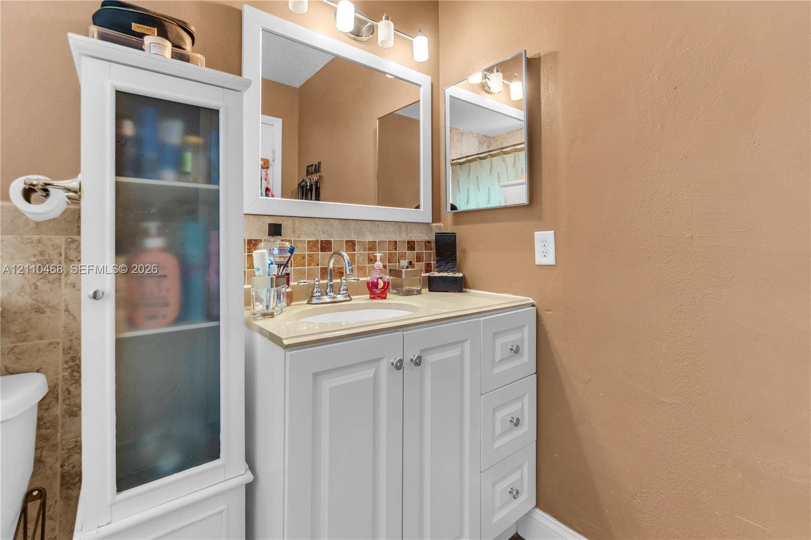 White vanity with stone-finish countertop, integrated sink, tiled backsplash, wall-mounted mirror, and attached glass-front cabinetry