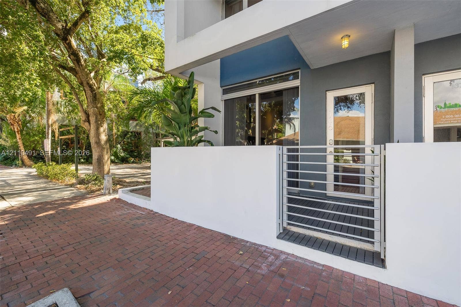 3339 Virginia Street, #136, Miami, Miami, FL 33133 Modern exterior entrance featuring a white stucco privacy wall, horizontal slat metal gate, red brick paver walkway, and recessed overhead lighting