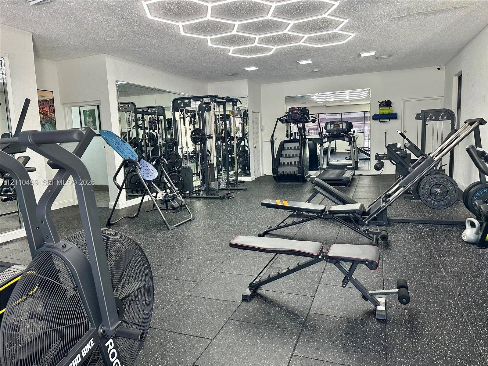 8145 NW 7th Street, #308, Miami, Miami, FL 33126 Fitness space featuring black rubberized flooring, large-scale wall-to-wall mirrors, and a custom hexagonal geometric LED ceiling light fixture