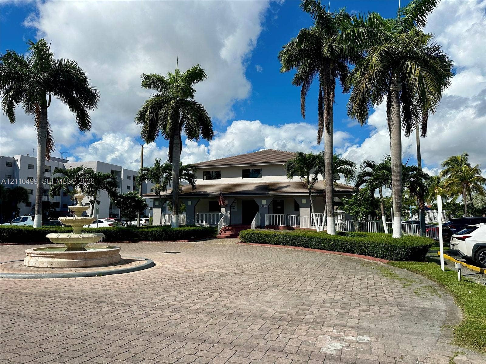 8145 NW 7th Street, #308, Miami, Miami, FL 33126 Paver brick circular driveway featuring a tiered stone fountain, manicured hedges, and mature palm trees