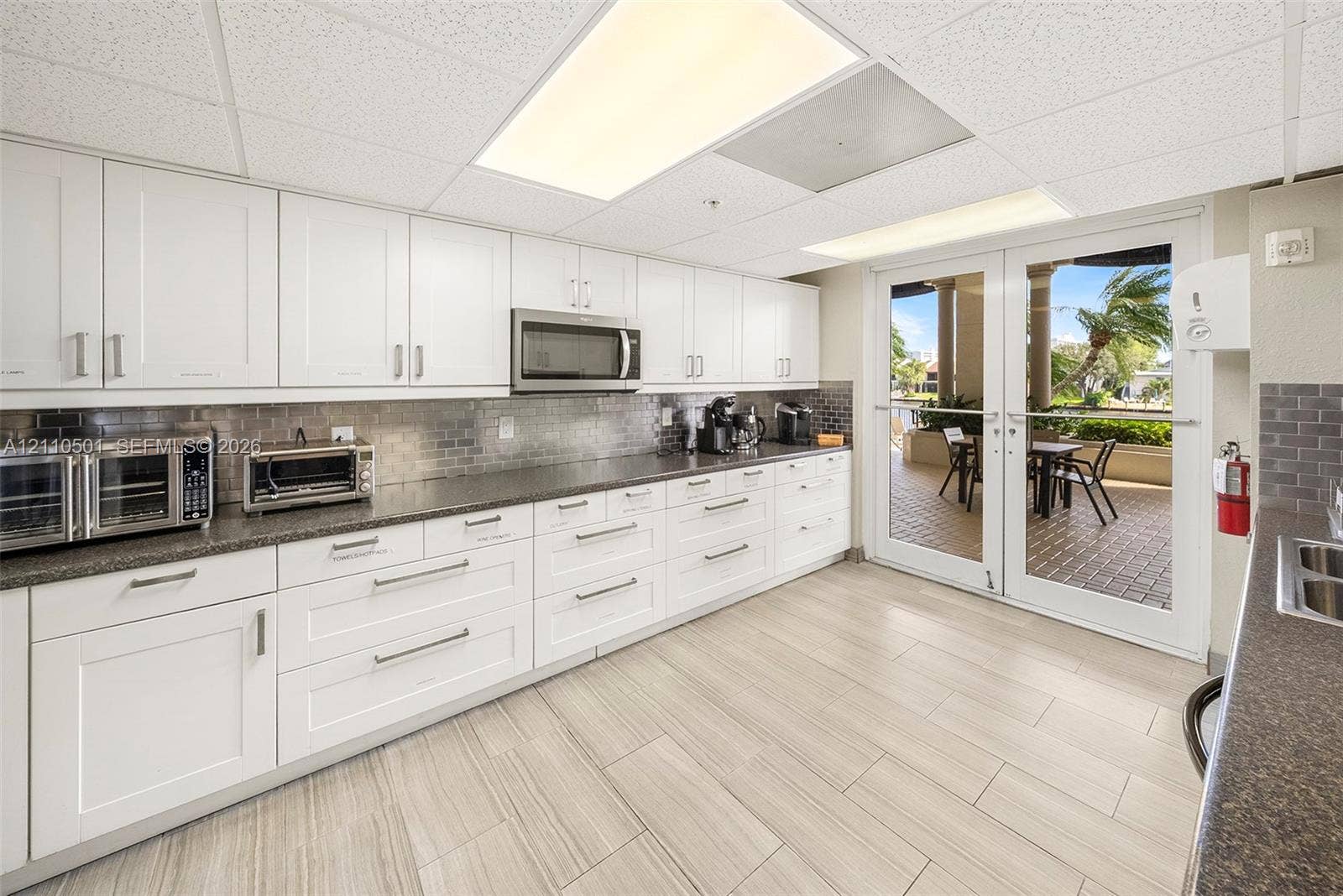 Kitchen featuring white shaker-style cabinetry, dark stone-finish countertops, subway tile backsplash, light-toned tile flooring, and double French doors opening to an outdoor patio