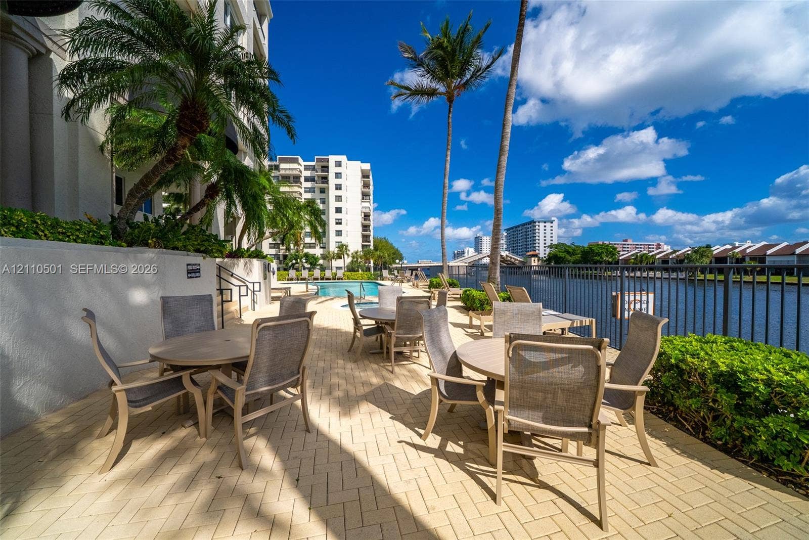 Waterfront patio featuring herringbone paver hardscaping, mature palm trees, and metal perimeter fencing