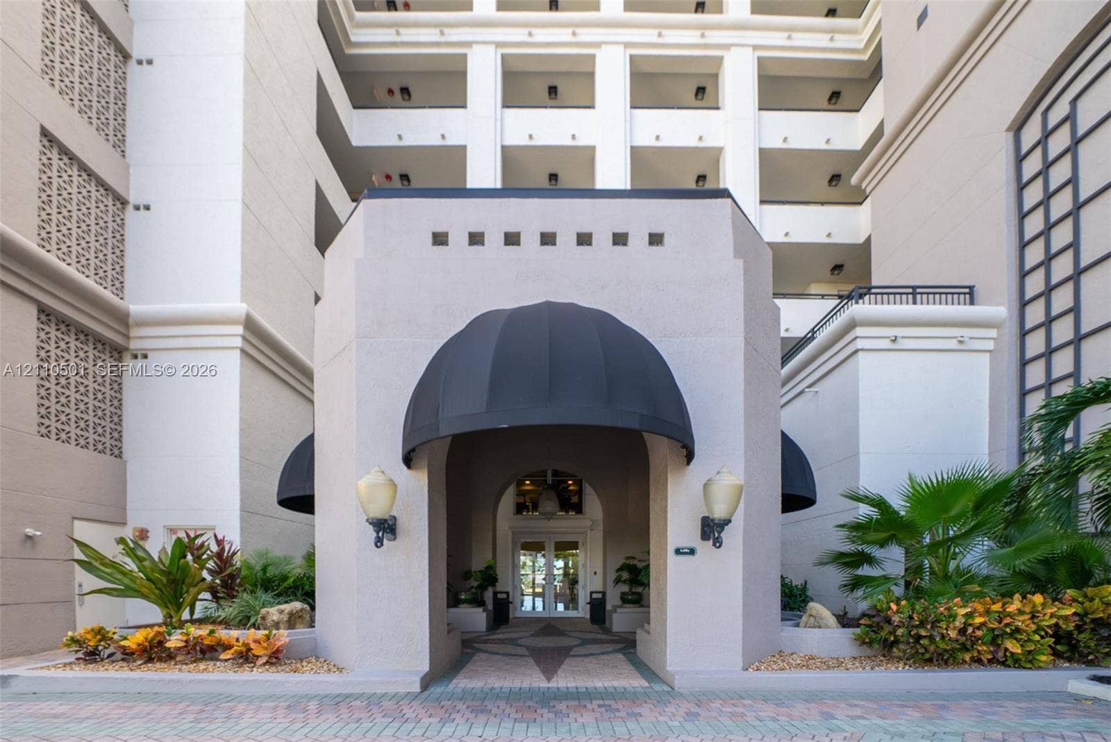 Stucco porte-cochere with dual black fabric awnings, decorative masonry inlay flooring, wall-mounted exterior globe lighting, and manicured landscaping