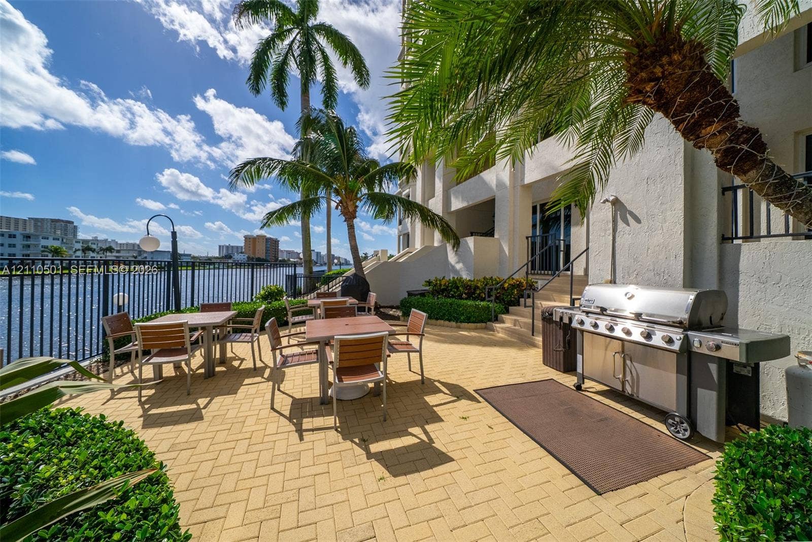 Patio featuring herringbone-patterned pavers, built-in stainless steel grill, metal fencing, and mature palm trees