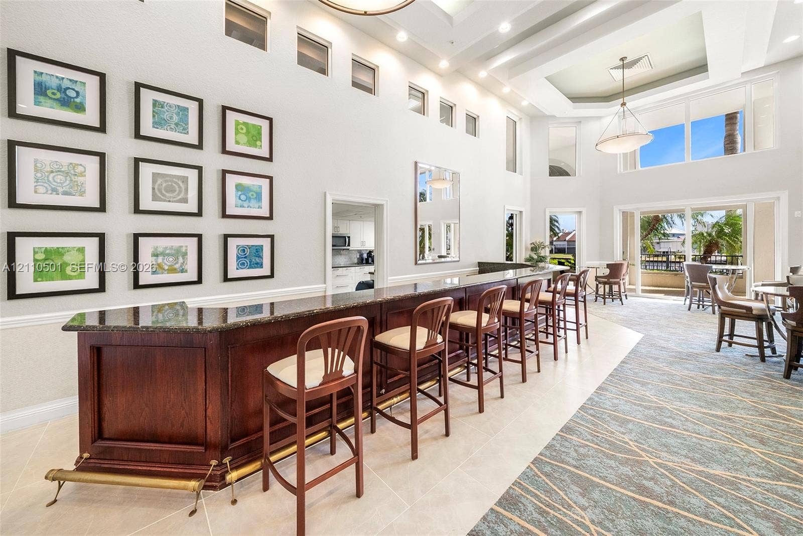Custom wet bar featuring dark wood cabinetry, stone countertops, and a brass-tone foot rail, set within a high-ceiling space accented by coffered ceilings, multi-level windows, and recessed lighting