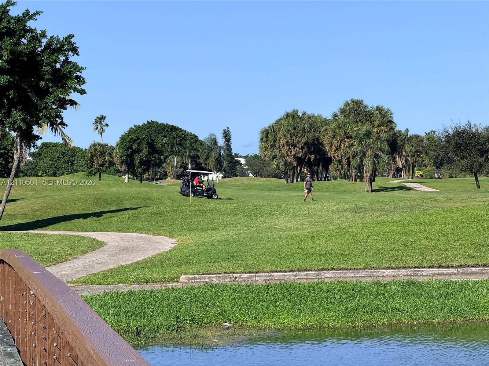 Golf course grounds featuring a paved pathway, a pedestrian bridge, a water feature, and mature palm trees