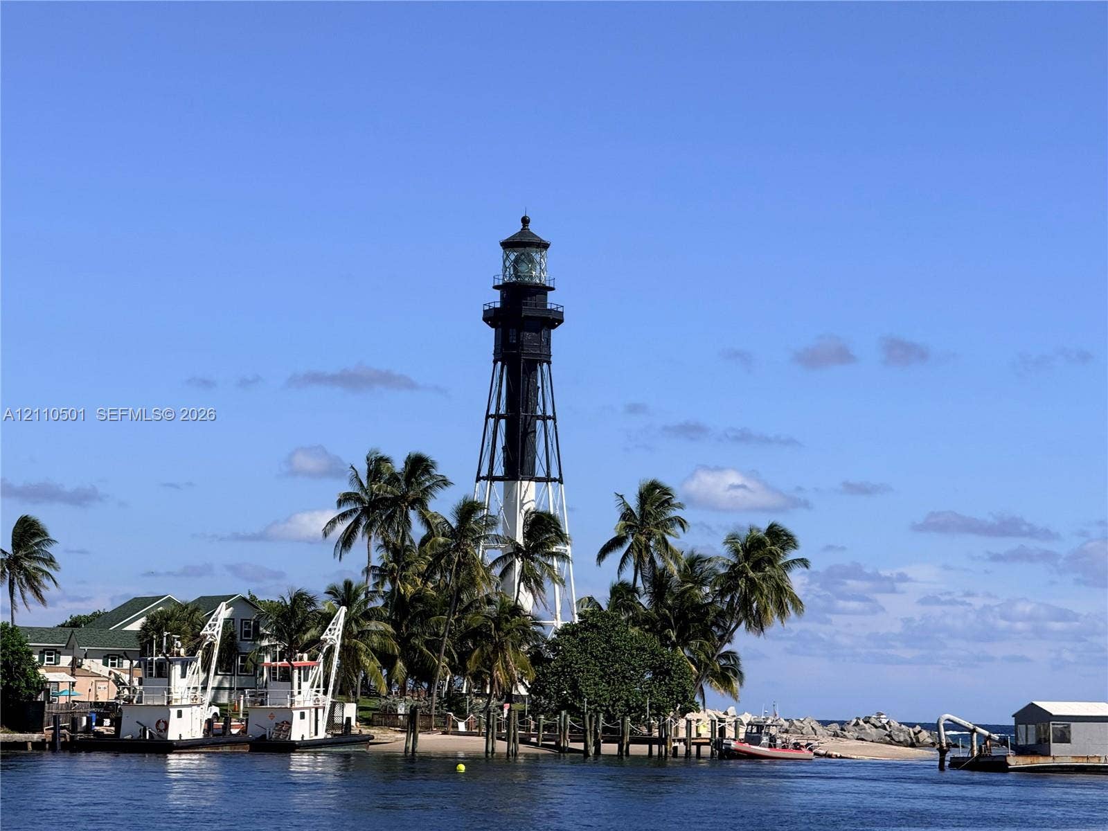 Lighthouse structure featuring a cylindrical metal tower design, a prominent gallery railing, and lantern room, surrounded by mature palm trees and coastal rock embankment
