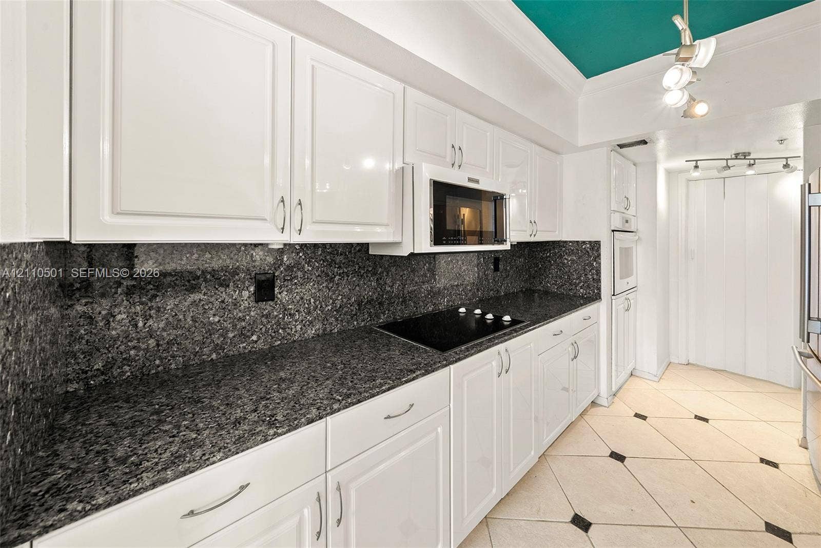 Kitchen featuring white cabinetry, stone countertops with a matching full-height backsplash, built-in cooktop, integrated microwave, and light-toned tile flooring with contrasting inlay accents