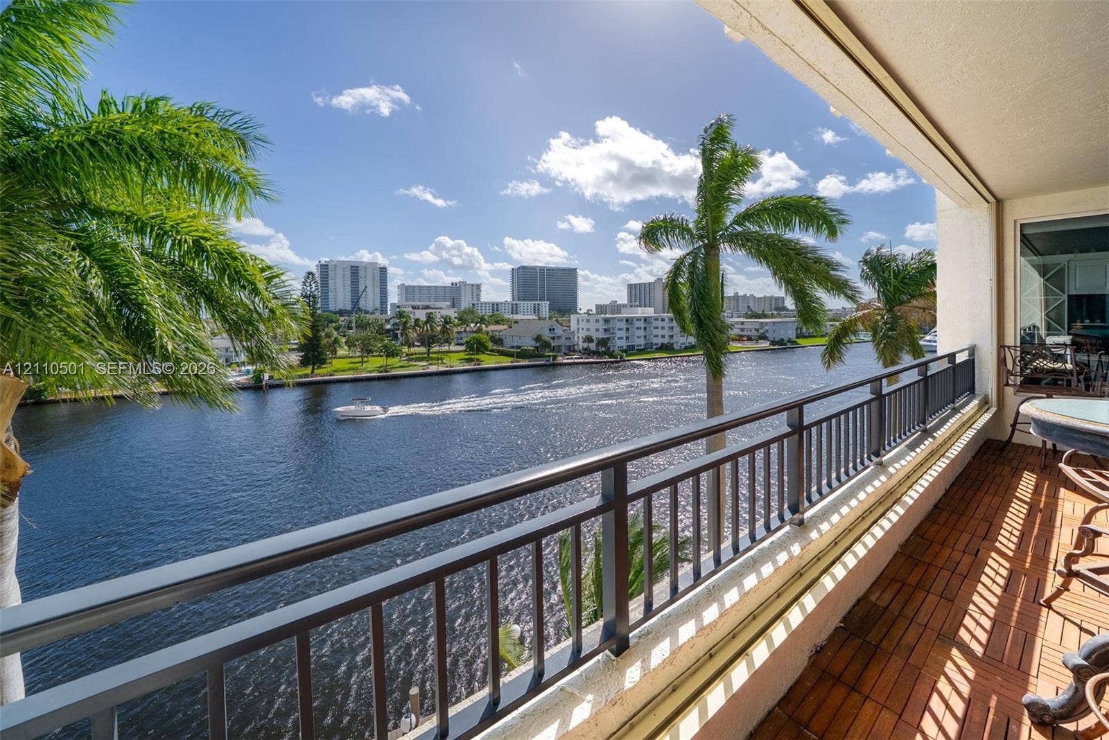 Covered balcony featuring wood-finish decking and a dark metal railing, overlooking a waterway with urban architecture in the background